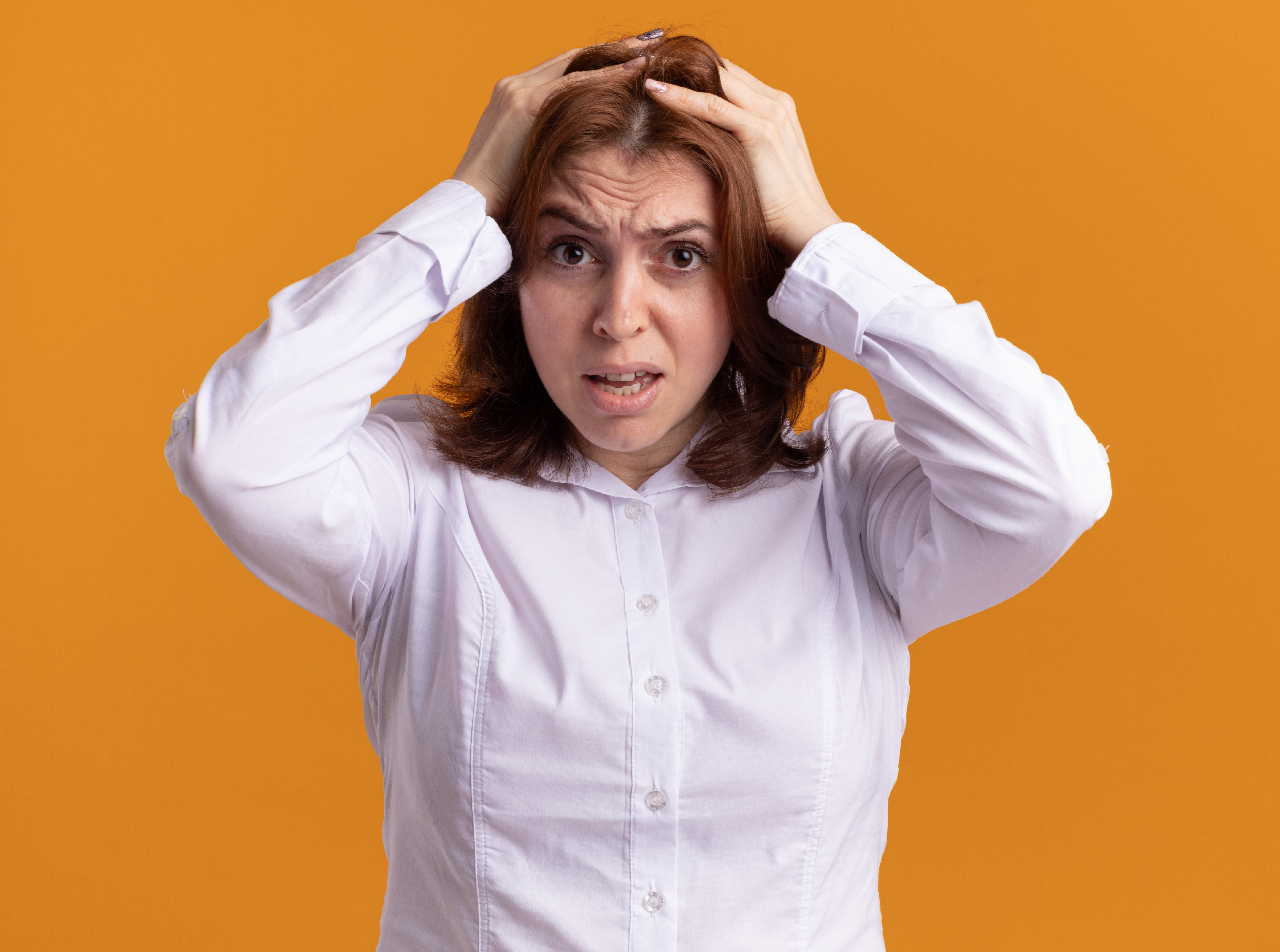young woman in white shirt lookign at camera confused and frustrated with hands on her head for mistake standing over orange background
